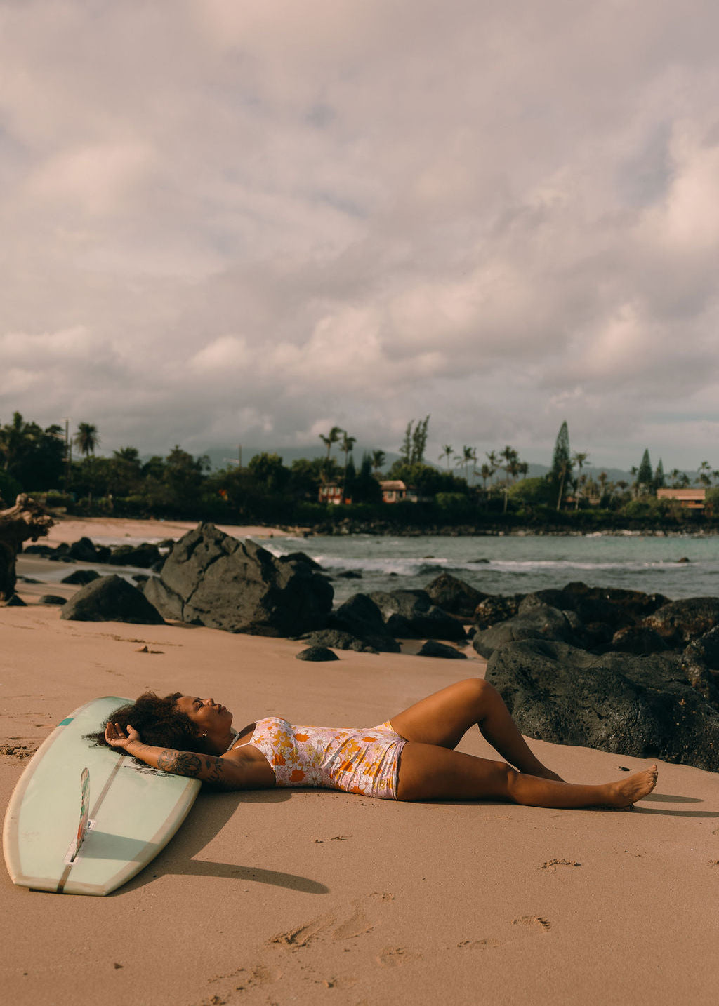 Woman in sustainable surf suit lying on a beach with a surfboard, surrounded by rocks and ocean.