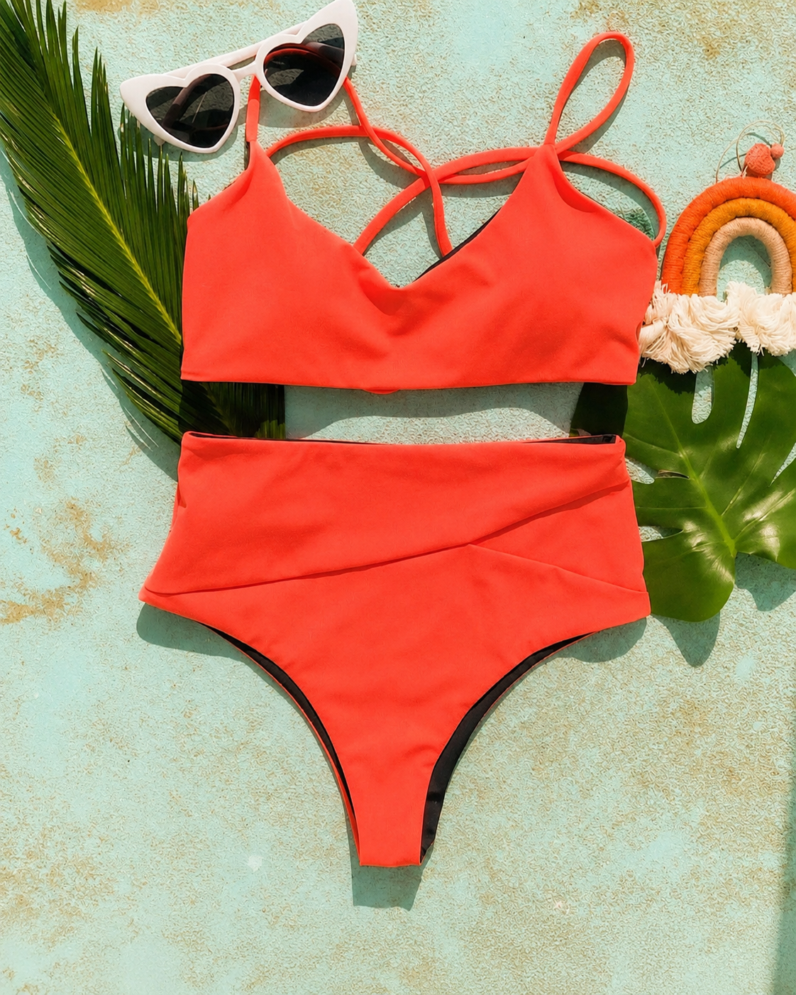 Flat-lay of a coral red bralette top and high-waist bikini bottoms with heart sunglasses and a palm leaf.