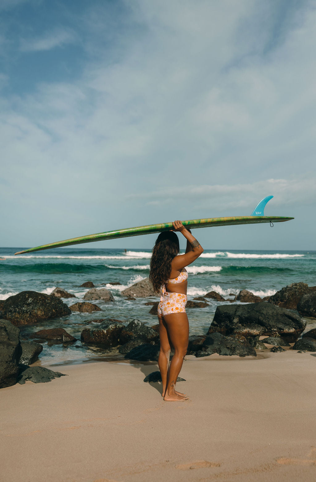 Woman carrying a surfboard on a beach with rocks and ocean waves in the background