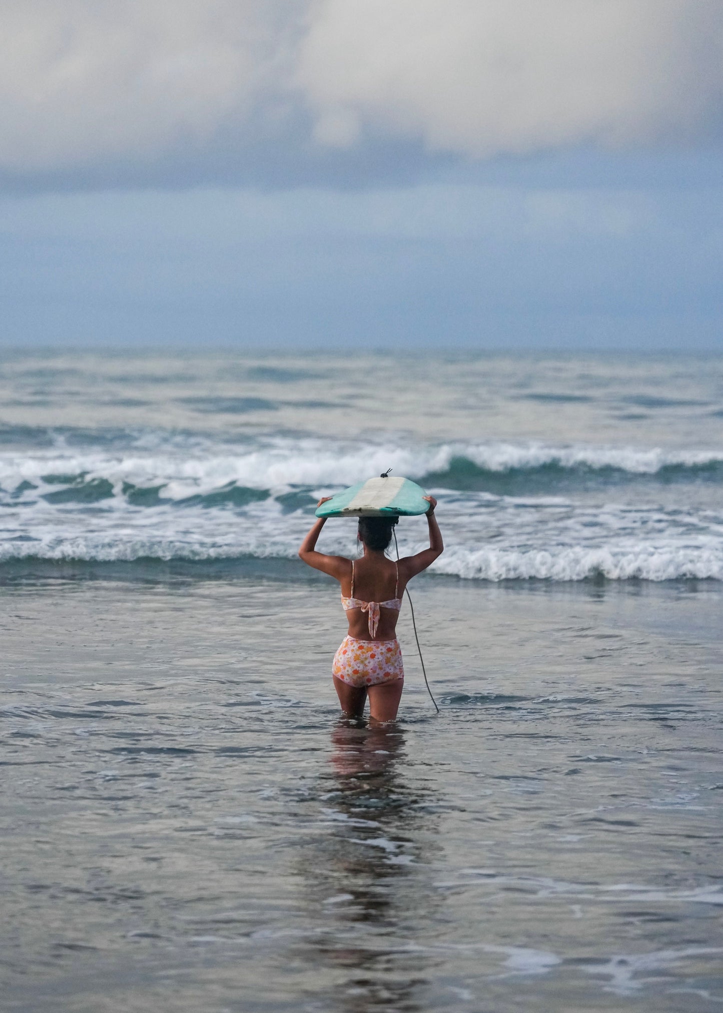 Surfer girl in purple floral bikini holding a surfboard above their head while standing in the ocean.
