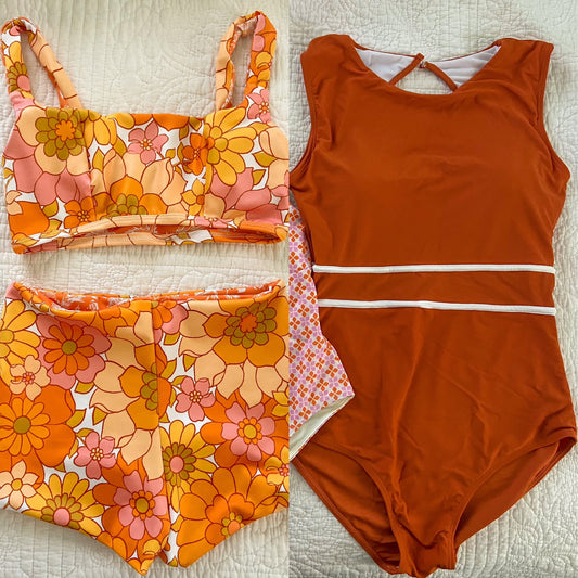 Woman walking a rocky beach holding a surfboard in an orange floral one-piece, the San O classic surf suit.